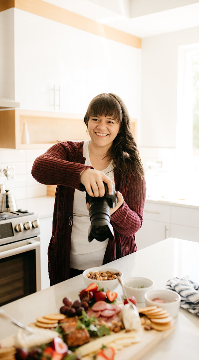 Smiling, long, brown-haired, woman in a red sweater and white shirt in a kitchen, holding camera over a cheese board.