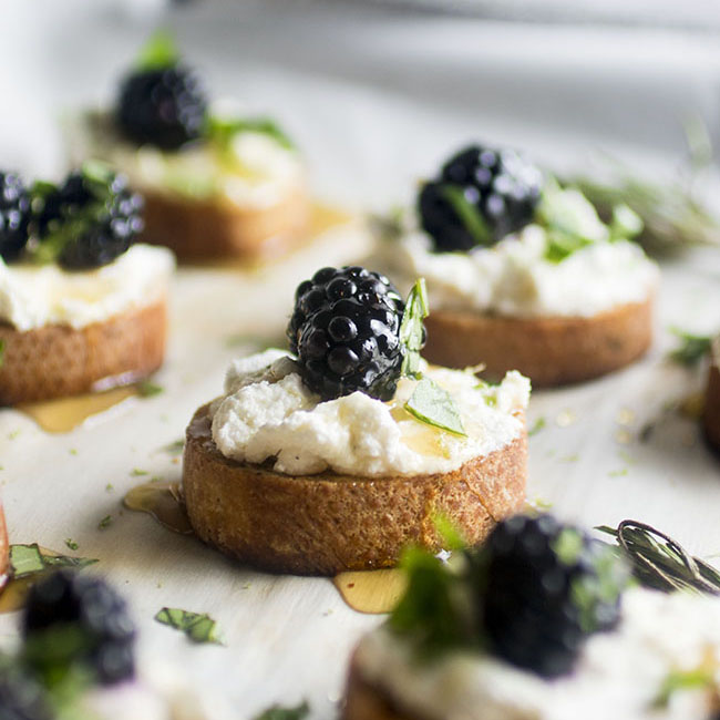 round pieces of toasted baguette on a sheet pan. Each is topped with white cheese, honey, blackberry and chopped basil
