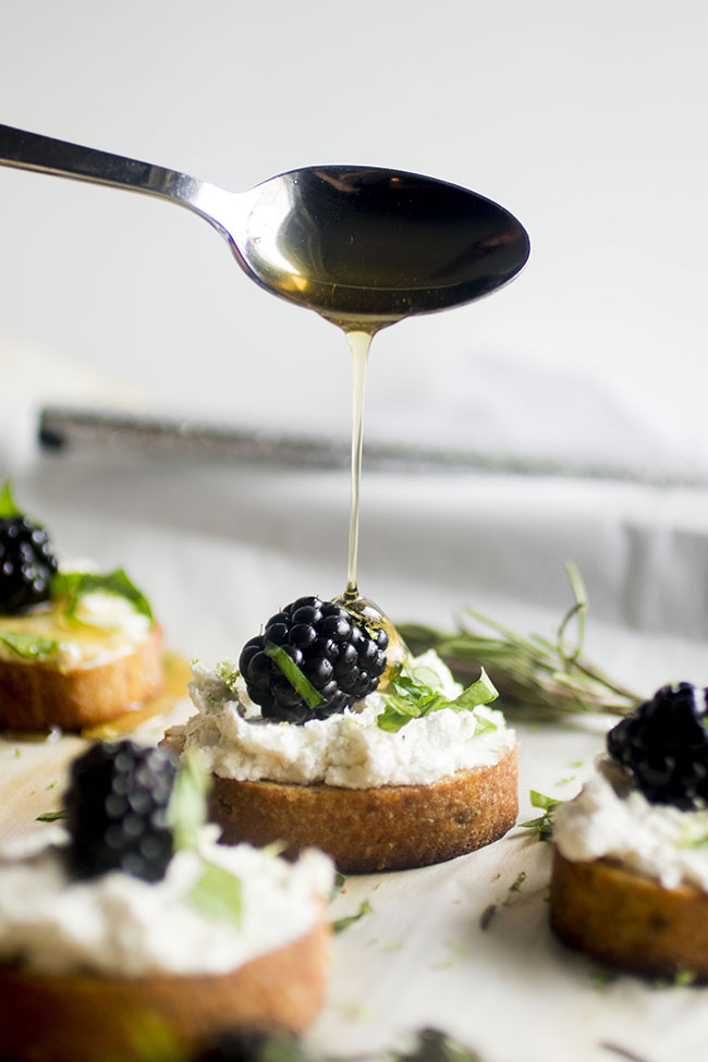 round pieces of sliced, toasted baguette topped with white goat cheese, a blackberry and chopped basil. A spoon is drizzling honey over the top of the blackberry and cheese.
