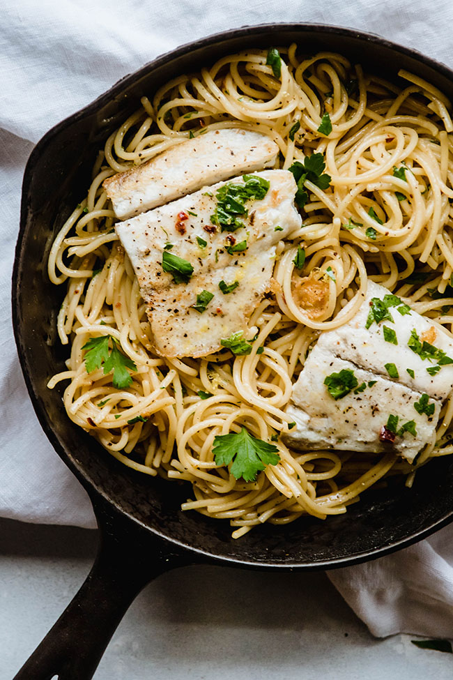 black cast iron skillet on a white cloth. In skillet is spaghetti noodles topped with barramundi fish fillets and cilantro
