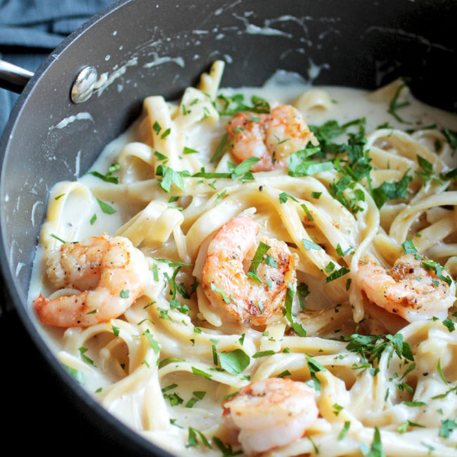 Black skillet with fettuccine noodles, prawns and green parsley in a white creamy, alfredo sauce.