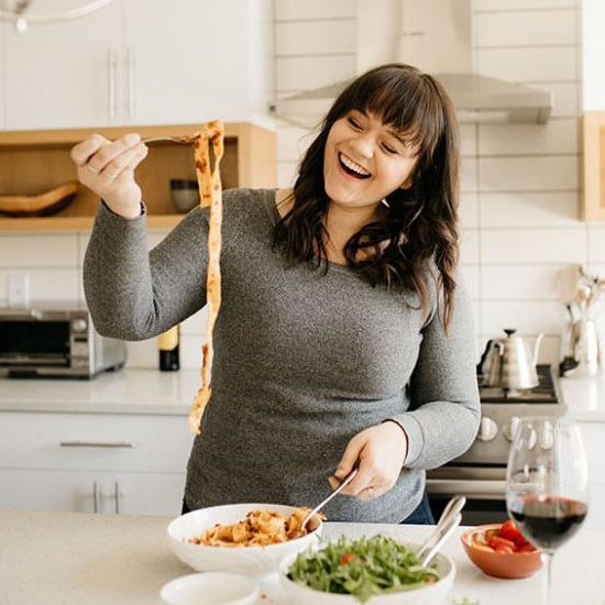 Long, dark-haired smiling woman in a kitchen. holding a long piece of pappardelle with marinara sauce on a fork over a bowl of pasta. On counter is bowl of pasta, arugula salad and glass of wine