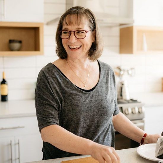Short, brown-haired, woman in a black and gray shirt laughing in the kitchen